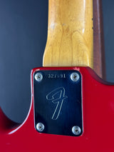 Close-up of the tailpiece area of a red electric guitar, showing a black metal plate with a white signature logo and serial number 327591, next to a natural wood neck.