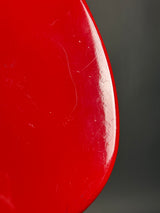 A close-up of a glossy red curved surface, likely part of a chair or stool, with visible scratches and scuffs on the red finish, set against a dark gray background.