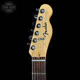 Close-up of the headstock of a Fender Telecaster electric guitar in natural wood, showing six silver tuning pegs and the black Fender logo, set against a solid black background.