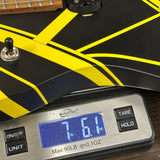 A digital scale displaying 7 lbs 6 oz is shown under a black and yellow electric guitar body on a dark surface.