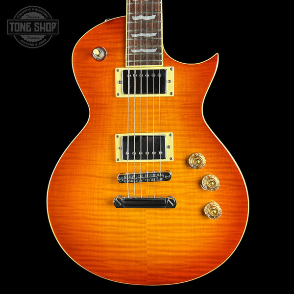 A close-up of an electric guitar body in a vibrant orange sunburst finish, featuring gold hardware, black pickups, and a dark fretboard, set against a solid black background.