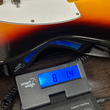 A close-up of a smart weigh scale displaying a weight of 81.4 kg under a sunburst electric guitar on a dark wooden surface.