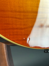Close-up of a glossy wood guitar body showing a warm orange-brown finish, a white binding edge, and part of a black hardware component against a dark gray background.