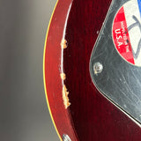 Close-up of a glossy dark red electric guitar body showing a broken edge finish, scratches on the black pickguard, and part of a red inspection sticker.