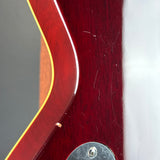 Close-up of a glossy dark red wooden guitar body with a gold outline edge, a silver metal hardware piece, and visible surface scratches on a white background.