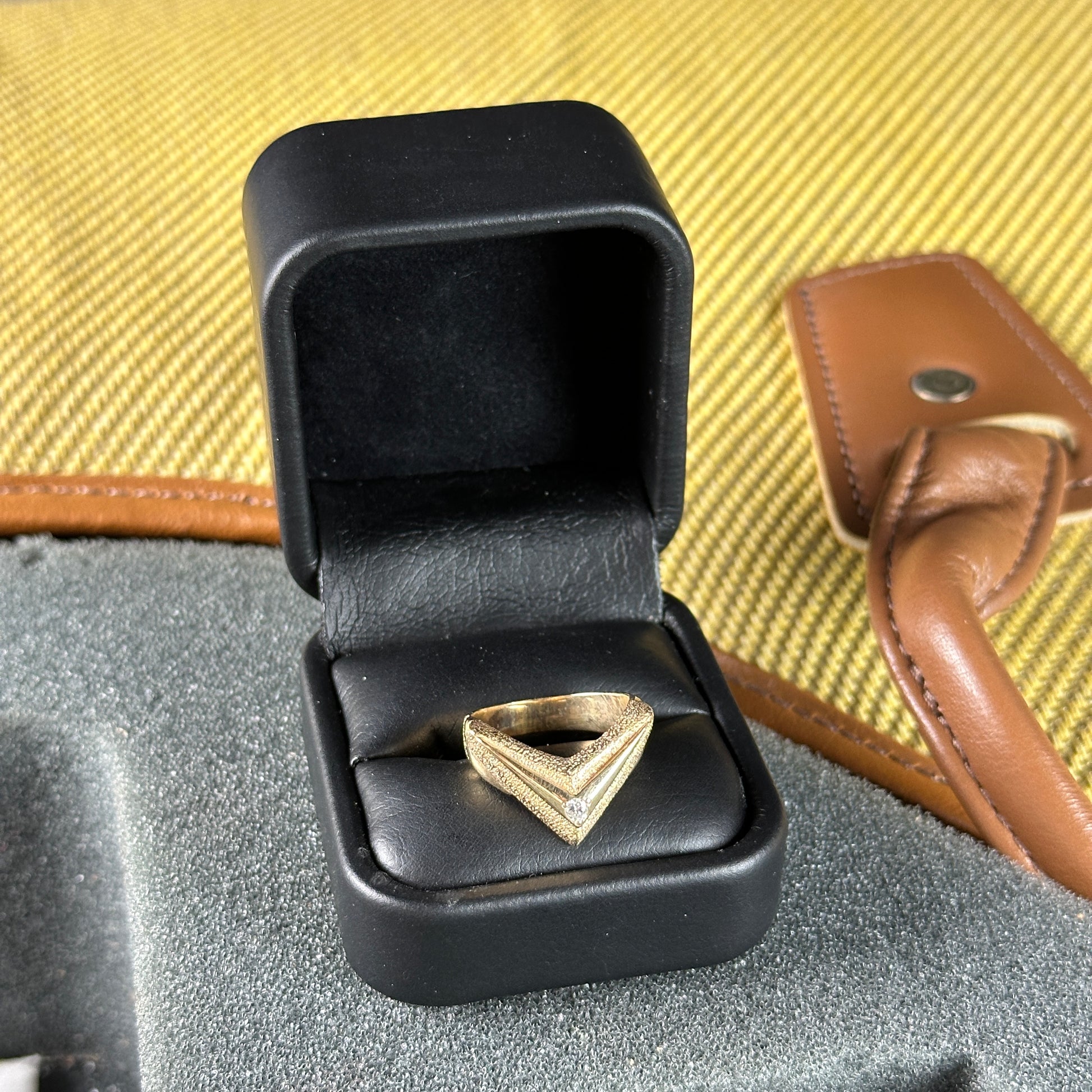 Gold triangular ring in a black jewelry box on a textured surface with a brown leather item in the background.