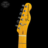 Close-up of a Fender Telecaster electric guitar headstock in a natural maple wood finish, featuring six silver tuning pegs and the black Fender logo, set against a solid black background.