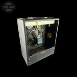 A vintage Fender amplifier cabinet with its front speaker grille removed, showing the internal speaker, wiring, and metal components. The cabinet is covered in textured gray speaker cloth with a silver handle on top.