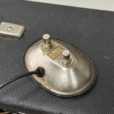 A close-up of a vintage metal dual-switch control plate with a black cable connected, set against a textured black surface.