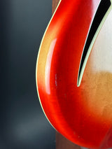 Close-up of a glossy sunburst electric guitar body showing the red-to-brown gradient finish and the black double-cutaway opening.