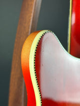 Close-up detail of a guitar body corner showing the glossy reddish-brown finish, cream binding, and light wood edge against a dark background.