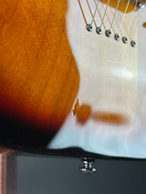 Close-up of a sunburst electric guitar body showing the glossy amber-brown wood finish, part of the white headstock, and a silver tuning peg.
