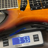 A close-up of a guitar body with a digital scale placed in front of it on a dark wooden surface. The scale reads 7.53 oz.