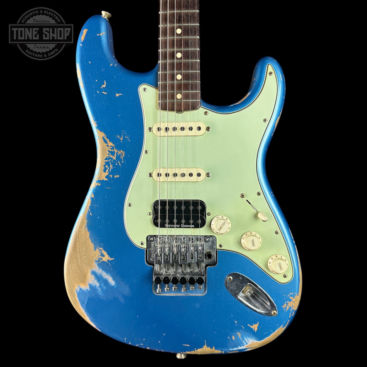 A blue electric guitar with a light green pickguard and distressed finish, shown against a black background with a Tone Shop watermark in the top left corner.