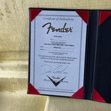A Fender Certificate of Authenticity document displayed inside a black folio with red corner accents, set against a textured gold fabric background.