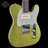 A close-up of a Fender Telecaster electric guitar body in a metallic gold glitter finish, featuring a light gray pickguard, dual humbucker pickups, and chrome hardware, set against a solid black background.