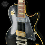 Close-up of a black electric guitar with gold hardware on a black background, featuring the 'Tone Shop' logo.