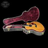 An acoustic guitar with a natural wood finish and dark pickguard displayed inside an open hard case with a burgundy velvet interior, set against a solid black background.