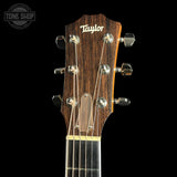 Close-up of a Taylor acoustic guitar headstock with six silver tuning pegs and six strings, set against a solid black background. A Tone Shop watermark is visible in the top left corner.