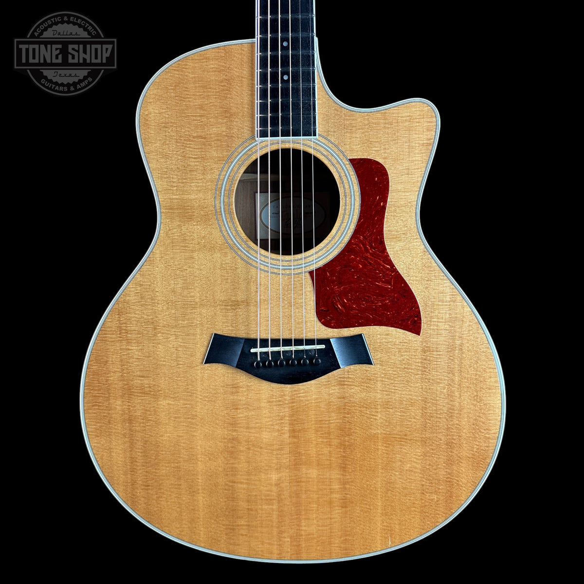 A Taylor acoustic guitar with a natural flamed maple top, dark fretboard, and a red tortoiseshell-style pickguard, shown against a solid black background with a Tone Shop watermark in the top left corner.