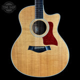 A Taylor acoustic guitar with a natural flamed maple top, dark fretboard, and a red tortoiseshell-style pickguard, shown against a solid black background with a Tone Shop watermark in the top left corner.