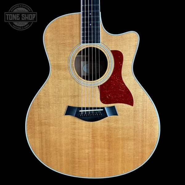 A Taylor acoustic guitar with a natural flamed maple top, dark fretboard, and a red tortoiseshell-style pickguard, shown against a solid black background with a Tone Shop watermark in the top left corner.