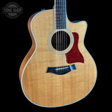 A Taylor acoustic-electric guitar with a natural wood finish, black cutaway body, and a red pickguard, shown against a solid black background with a Tone Shop watermark in the top left corner.