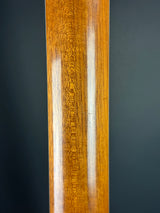 A close-up of a polished wooden rail or trim piece with a warm golden-brown wood grain finish, showing a glossy reflection, set against a dark gray background.