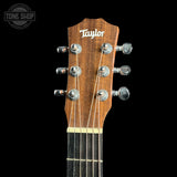 Close-up of a Taylor acoustic guitar headstock with six silver tuning pegs and six strings, set against a solid black background. A Tone Shop watermark is visible in the top-left corner.
