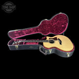 An acoustic-electric guitar with a natural wood body and dark neck, displayed inside an open hard case with a deep magenta velvet lining, set against a solid black background.