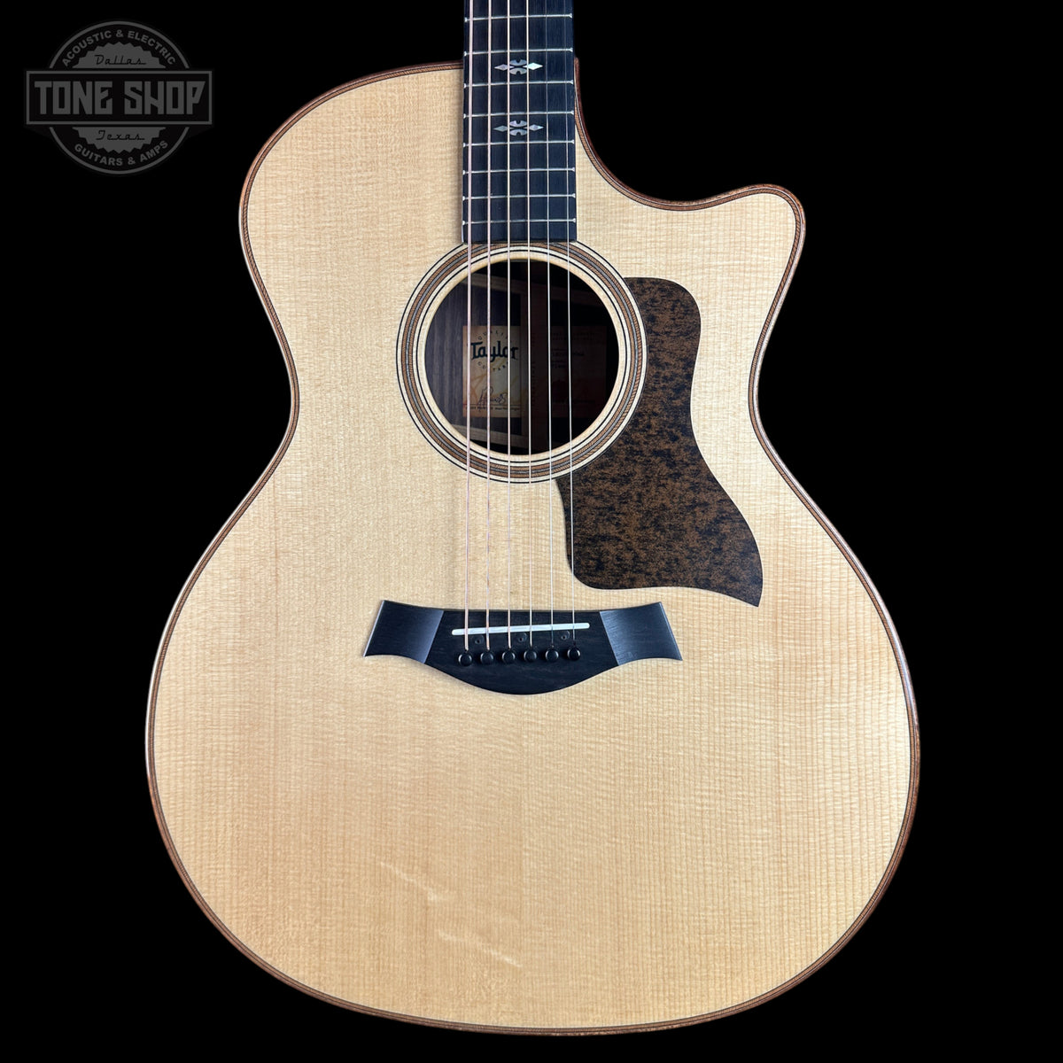 A Taylor acoustic-electric guitar with a natural wood finish and dark pickguard, shown against a solid black background with a Tone Shop watermark in the top left corner.