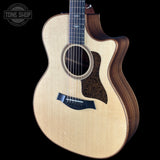 A Taylor acoustic-electric guitar with a natural wood top and dark wood sides, shown against a solid black background. A 'TONE SHOP' watermark is visible in the top-left corner.