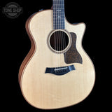 A Taylor acoustic-electric guitar with a natural wood finish and dark pickguard, shown against a solid black background with a Tone Shop watermark in the top left corner.