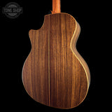 The back of an acoustic guitar with a rich dark brown wood grain finish and a light natural wood edge, shown against a solid black background with a Tone Shop watermark in the top left.