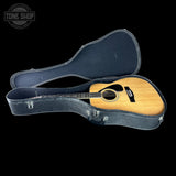 An acoustic guitar with a natural wood finish resting inside an open black hard case on a black background. A Tone Shop watermark is visible in the top-left corner.