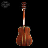 The back of an acoustic guitar with a glossy reddish-brown wood finish and a gold-toned center stripe, shown against a solid black background. A Tone Shop watermark is visible in the top-left corner.