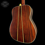 The back of an acoustic guitar with a rich dark brown wood grain finish, featuring two gold-toned binding strips and a yellow headstock visible at the top, set against a solid black background with a Tone Shop watermark in the upper left.