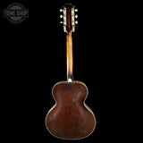 The back of a vintage acoustic guitar with a dark brown wooden body and neck, shown against a solid black background. A circular 'TONE SHOP' watermark is visible in the top left corner.