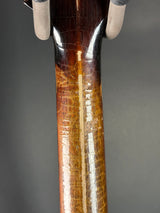 Close-up of a polished wooden gun stock with a glossy finish, showing natural wood grain and minor wear, held by hands wearing light-colored gloves against a dark gray background.