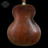 The back of a vintage acoustic guitar with a glossy dark brown wooden body, showing natural wear and scratches, set against a solid black background with a Tone Shop watermark in the top left.