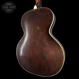 The back of a vintage acoustic guitar with a dark brown varnished wooden body and a cream-colored binding edge, shown against a solid black background.