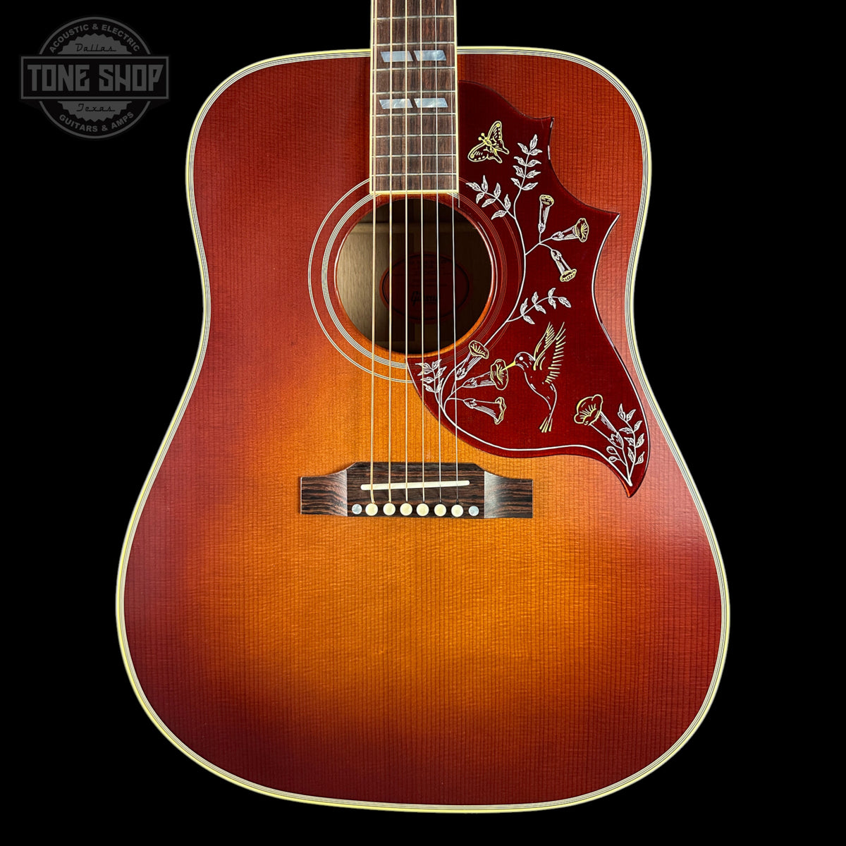 A close-up of an acoustic guitar body against a black background, featuring a natural sunburst finish that transitions from bright orange in the center to dark brown at the edges, with a red pickguard decorated with white floral and bird inlays.