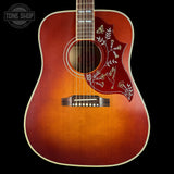 A close-up of an acoustic guitar body against a black background, featuring a natural sunburst finish that transitions from bright orange in the center to dark brown at the edges, with a red pickguard decorated with white floral and bird inlays.