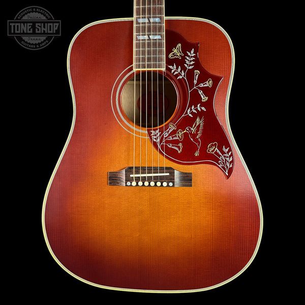 A close-up of an acoustic guitar body against a black background, featuring a natural sunburst finish that transitions from bright orange in the center to dark brown at the edges, with a red pickguard decorated with white floral and bird inlays.