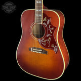 A classic acoustic guitar with a warm brown sunburst finish, featuring white floral inlay on the pickguard and a white binding around the body, shown against a solid black background.