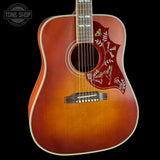 A close-up of an acoustic guitar body with a natural sunburst finish, featuring a red pickguard with gold floral and bird inlays, set against a solid black background.
