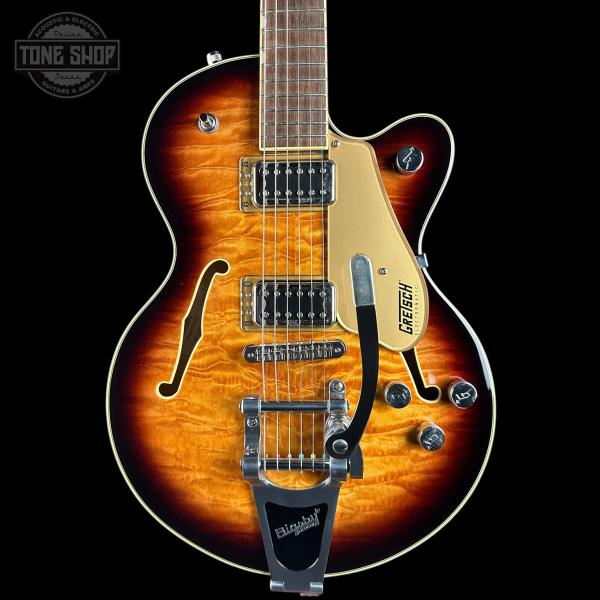 A Gretsch double-cutaway electric guitar with a natural flame maple top, shown from the front against a black background. The guitar features a sunburst finish transitioning from amber-orange to dark brown, with a cream pickguard, chrome hardware, and a black tremolo system.