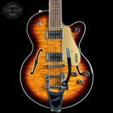 A Gretsch double-cutaway electric guitar with a natural flame maple top, shown from the front against a black background. The guitar features a sunburst finish transitioning from amber-orange to dark brown, with a cream pickguard, chrome hardware, and a black tremolo system.
