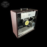 A Fender Custom Shop '73 Bass amplifier head shown from a three-quarters angle against a solid black background, revealing the internal speaker, heatsink, and wiring. Features a brown textured exterior and a white control panel on the back.