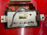 A close-up of a used guitar humbucker pickup mounted on a red guitar body. The pickup has a silver metal faceplate with brass screws, a black surround, and a braided cable.